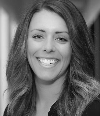 Professional headshot of Natalie Toth, a woman with long wavy hair, smiling brightly at the camera, wearing a dark top against a softly blurred indoor background, in black and white