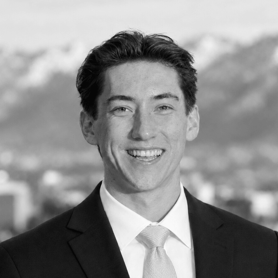 Black and white headshot of a smiling man in a dark suit and tie, with a blurred mountain and city skyline in the background